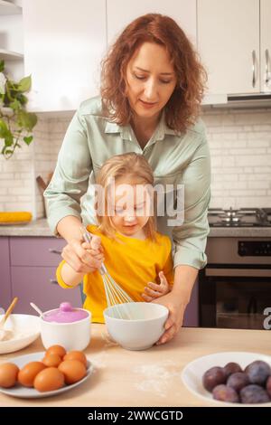 Mutter und Tochter kochen Kuchen in lila Küche, Training und Entwicklung, interessantes Spiel und Hilfe. Glückliche Familie zusammen. Stockfoto