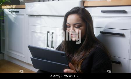 Asiatische Frau sitzt auf dem Boden in der Küche und genießt mit einem Tablet-Gerät im Internet surfen, Spiele spielen, im Internet einkaufen, Social Media Apps nutzen Stockfoto