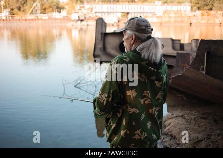 Der ältere Mann fischt am Fluss. Schmutziges Wasser, Verschmutzung, Müll, rostige Binnenschiffe. Ökologische Katastrophe, Produktionsabfälle. Stockfoto