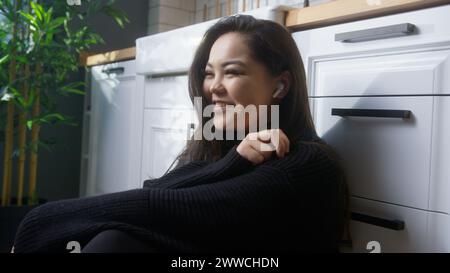 Lächelnde asiatische Mädchen sitzen auf dem Boden in der Küche und hören Musik mit kabellosen in-Ear-Kopfhörern. Eine Frau genießt es, Zeit allein zu Hause zu verbringen Stockfoto