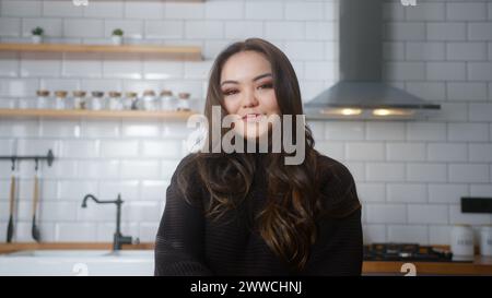 Die junge asiatin sitzt in der Küche und sieht in die Kamera und lächelt. Gemütliche Frau, die sich im Haus entspannt Stockfoto