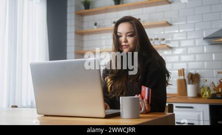 Asiatische Frau, die Kreditkartennummer auf dem Laptop für Online-Shopping eingeben, macht sichere, einfache elektronische Zahlung in entfernter Entfernung, während Sie in der Küche zu Hause sitzen Stockfoto