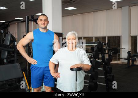 Eine Rentnerin, eine ältere Frau, führt eine Übung mit einer Langhantel im Fitnessstudio aus. Der Coach hilft und zeigt. Gesunder Sportstil, Seniorenkonzept. Frau mit Stockfoto