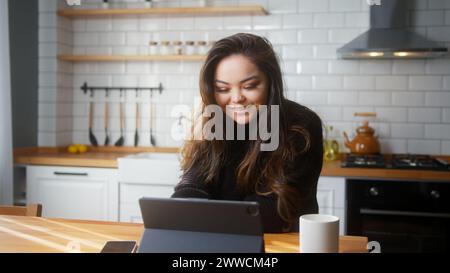 Asiatisches Mädchen sitzt in der Küche und führt Videogespräche über einen Tablet-Computer. Teilen Sie Neuigkeiten, sprechen Sie mit der Familie, genießen Sie virtuelle Webchat-Meetings Stockfoto