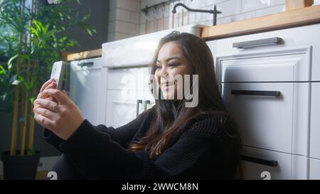 Lächelnde asiatische Frau sitzt auf dem Boden in der Küche und hält ihr Handy, winkt und grüßt, während sie über Videokonferenzgespräche auf dem Smartphone spricht. Frau spricht, smi Stockfoto
