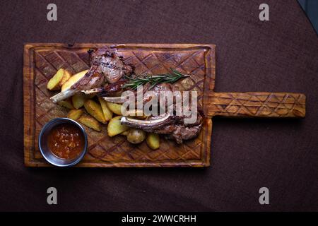 Zwei saftige Lammkoteletts und knusprige Kartoffelscheiben serviert auf einem rustikalen Holzbrett mit einer Beilage aus würziger Dippsoße. Stockfoto