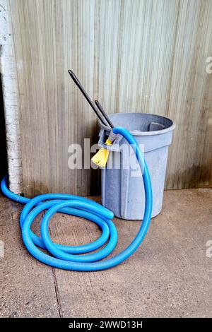 Langer blauer Schlauch mit gelber Düse, eingehakt auf grauem Abfalleimer Stockfoto