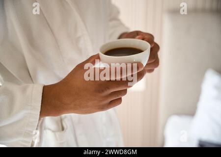 Nahaufnahme einer Tasse Kaffee in den Händen eines Mannes mit Bademantel im Hotelzimmer Stockfoto