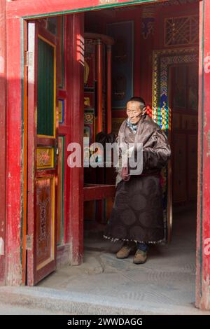 Ältere männliche Pilger, Gesicht im Sonnenlicht gefangen, verlassen einen Raum mit einem großen Gebetsrad im Kloster Labrang, County Xiahe, Provinz Gansu, China Stockfoto