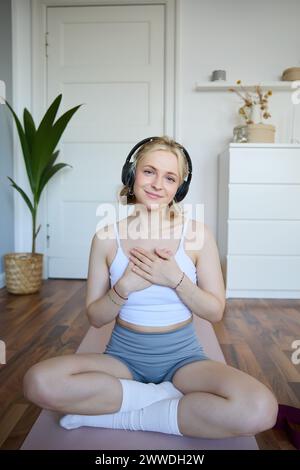 Vertikales Porträt einer schönen jungen Frau in Kopfhörern, sitzt auf Yogamatte, meditiert, hält die Hände auf der Brust und lächelt in die Kamera Stockfoto