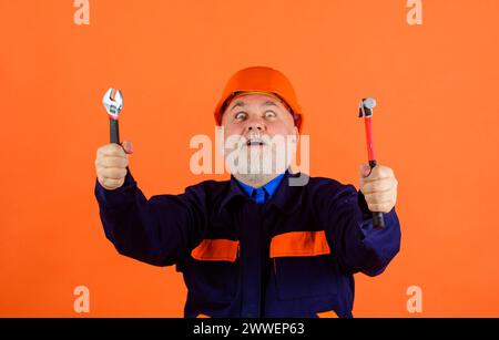 Überraschter männlicher Baumeister in Schutzhelm mit verstellbarem Schraubenschlüssel und Hammer. Mechaniker oder Reparaturarbeiter in Uniform und Bauhelm Stockfoto