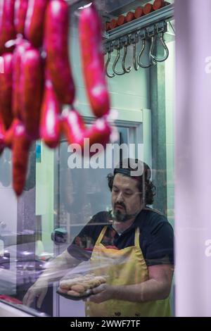 Vertikales Porträt eines Metzgers, der ein Tablett mit frisch zubereiteten Hähnchennuggets hält, durch das Fenster seiner Metzgerei gesehen. Im Vordergrund mehrere Stockfoto