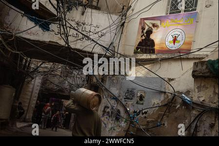 Beirut, Libanon. März 2024. Ein Mann trägt einen Gaskanister am 19. März 2024 im Shatilla Refugee Camp in Beirut, Libanon, an einem Plakat des islamischen Jihad vorbei. Das Shatilla Refugee Camp beherbergt etwa 30.000 Vertriebene Syrer und Palästinenser. (Foto: Collin Mayfield/SIPA USA) Credit: SIPA USA/Alamy Live News Stockfoto