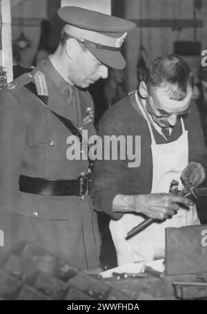 Ein Foto zeigt König Georg VI., der Interesse an der Arbeit eines der vielen Handwerker während des Zweiten Weltkriegs zeigt. Dieses Bild, das um 1940 aufgenommen wurde, zeigt das Engagement und die Unterstützung des Königs für die qualifizierten Arbeiter, deren Beiträge für die Kriegsanstrengungen von entscheidender Bedeutung waren, und spiegelt die Anerkennung des Monarchen für die Bedeutung aller Rollen im kollektiven Bemühen um den Sieg wider. Stockfoto