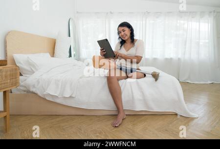 Eine Frau, die im Bett sitzt und in ein digitales Tablet vertieft ist. Eine prothetische bionische Beinprothese liegt auf ihrem Schoß. Stockfoto