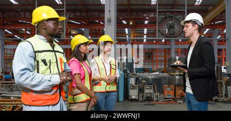 Gruppe von Ingenieur in Sicherheitsweste trifft sich mit einem jungen Manager in schwarzem Anzug in einer Industrie- oder Fabrikumgebung. Jeder trägt Schutzhelm. Bei Stockfoto