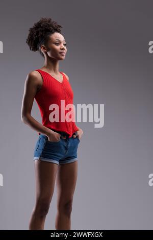 Die statueske Pose fängt das ruhige Selbstbewusstsein der Frau ein, gekleidet in Rot und Denim vor einem neutralen Hintergrund Stockfoto