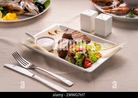 Gegrillte Würstchen oder Kupaty werden mit Gemüse, Salat und Ketchup auf einem weißen Teller serviert. Grillmenükonzept Stockfoto