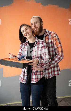 Lächelndes, liebevolles Paar, das Hausrenovierungen macht, Frau, die Tablett und Malerwalze hält, Mann umarmt sie Stockfoto