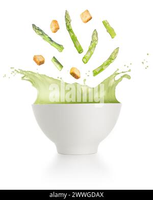 Spargel und Croutons fallen in eine cremige Suppenschüssel, die auf weißem Hintergrund spritzt. Stockfoto