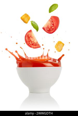 Tomaten, Croutons und Basilikumblätter, die in eine spritzende Schüssel Suppe fallen, isoliert auf weißem Hintergrund. Stockfoto