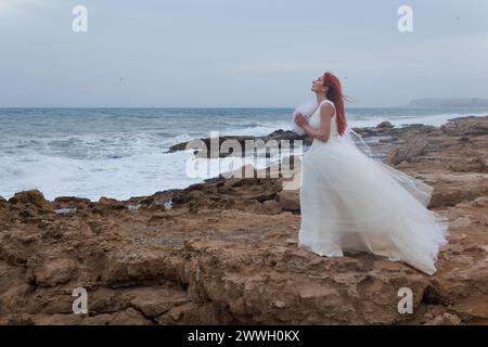 Einsame Frau im Hochzeitskleid betet zu Göttern ein Bräutigam für die Hochzeit an windigem Tag an der Mittelmeerküste, Alicante, Spanien Stockfoto