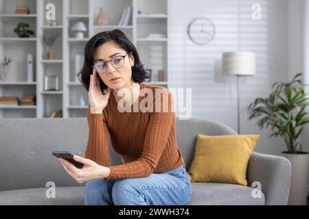 Eine Frau sitzt zu Hause auf ihrer Couch und schaut ihr Handy mit einem verwirrten Ausdruck an, möglicherweise nachdem sie schlechte Nachrichten erhalten hat. Stockfoto