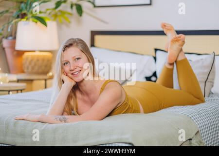 Eine entspannte junge Frau mit einem warmen Lächeln liegt auf einem Bett, bequem in legere Kleidung gekleidet, die ein Gefühl von häuslichem Komfort und Freizeit weckt. Stockfoto