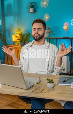 Friedlicher, entspannter männlicher Geschäftsmann, der am Arbeitsplatz mit gefalteten Mudra-Händen sitzt, Stress während des Arbeitstages reduziert, meditiert oder Yoga-Atemübungen im Heimbüro macht. Vertikal Stockfoto