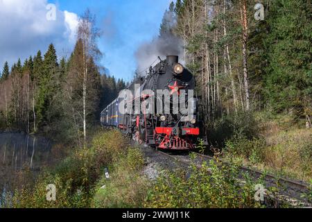 KARELIEN, RUSSLAND - 09. OKTOBER 2022: Eine alte Dampflokomotive mit Personenzug auf einem Waldstück in Karelien. Russland Stockfoto