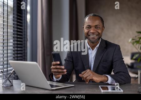 Porträt eines fröhlichen, reiferen afroamerikanischen Bosses im Business-Anzug, eines Geschäftsmannes, der lächelnd in die Kamera blickt und das Telefon in den Händen hält, eines Mannes, der im Büro mit einem Laptop arbeitet und die App verwendet. Stockfoto