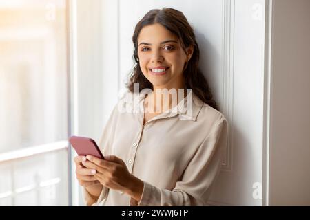 Glückliche junge Frau, die ein Smartphone hält und lächelt, während sie am Fenster steht Stockfoto