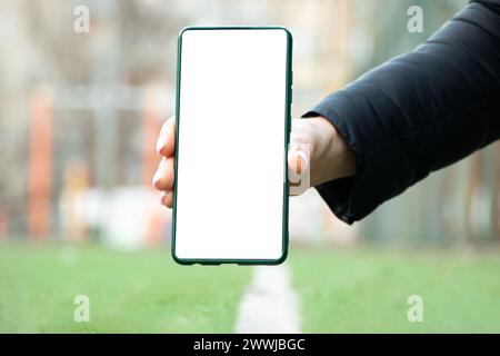 Die Hand einer Frau hält ein Telefon mit einem weißen Bildschirm auf einem Fußballfeld neben einem weißen Streifen, ein Mobiltelefon mit einem leeren Bildschirm Stockfoto