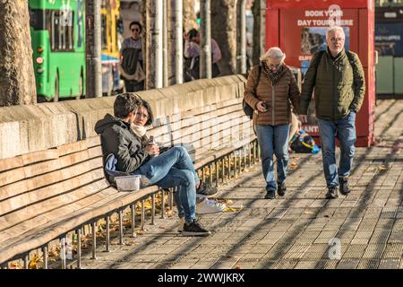 Jung und alt. Zwei Paare in derselben Aufnahme an einem sonnigen Tag. Das ältere Paar geht vorwärts, während das jüngere Paar sich auf einer Bank am Fluss entspannt. Stockfoto