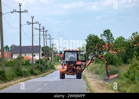 KINDER, SERBIEN - 5. JUNI. 2023: Traktor mit mechanischem Mäher, der Gras seitlich auf der Asphaltstraße mäht. Stockfoto