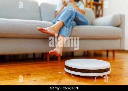 Person, die sich mit den Füßen nach oben auf dem Sofa entspannt, während der Roboter-Staubsauger auf dem Boden arbeitet Stockfoto