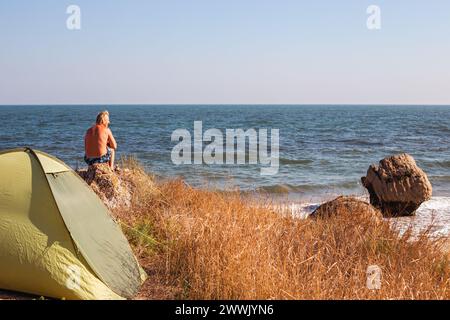 Reifer Mann sitzt an einer felsigen Küste neben einem Touristenzelt. Reisen und Tourismus. Stockfoto