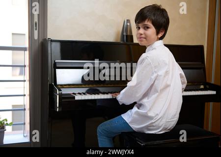 Selbstbewusster Teenager spielt zu Hause Klavier forte. Authentisches Porträt eines fröhlichen Teenager-Musikers in weißem Hemd, sitzt am Klavier und s Stockfoto