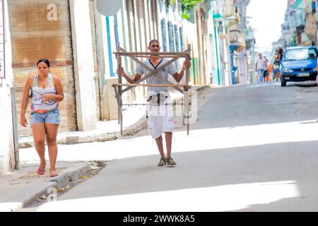 Ein kubanischer Mann trägt ein hölzernes Objekt mitten auf der Straße in Havanna, Kuba, während eine Frau auf dem Bürgersteig entlang läuft. Stockfoto