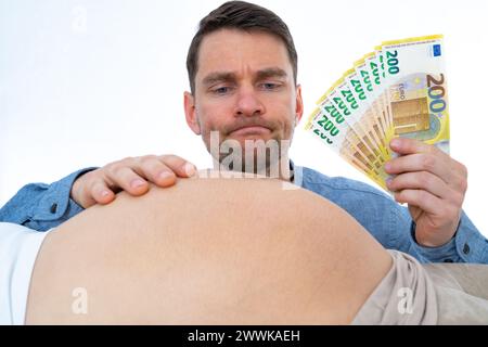 Beschreibung: Werdender Vater mit Banknoten in der Hand hält nachdenklich schwangere Babystöcke seiner Frau. Letzter Schwangerschaftsmonat - Woche 39. Weißer Hintergrund Stockfoto