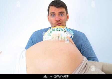 Beschreibung: Werdender Vater hinter schwangeren Babybauch hält einen Stapel Banknoten vor seinem Gesicht und sieht unsicher aus. Letzter Schwangerschaftsmonat Stockfoto