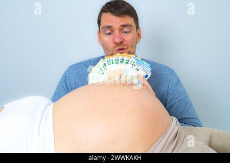 Beschreibung: Der werdende Vater hinter dem Bauch eines schwangeren Babys hält einen Stapel Banknoten vor seinem Gesicht und sieht besorgt aus. Letzter Schwangerschaftsmonat Stockfoto