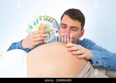 Beschreibung: Werdender Vater mit Banknotenstapel in der Hand hält nachdenklich schwangere Babystöcke seiner Frau. Letzter Schwangerschaftsmonat - Woche 39. Wh Stockfoto