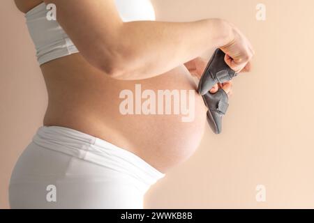 Beschreibung: Mittelteil einer nicht erkennbaren stehenden Mutter in weißen Tüchern, die Babyschuhe über ihren runden, schwangeren Bauch geht. Letzter Schwangerschaftsmonat - Stockfoto