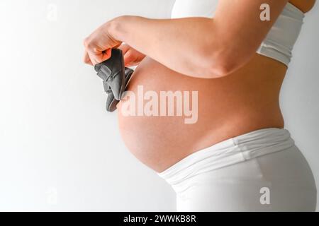 Beschreibung: Mittelteil einer nicht erkennbaren stehenden Mutter in weißen Tüchern, die Babyschuhe über ihren runden, schwangeren Bauch geht. Letzter Schwangerschaftsmonat - Stockfoto