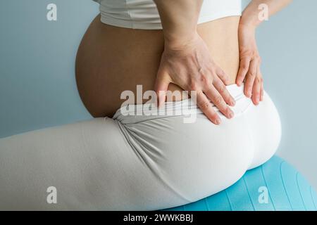 Beschreibung: Mittelteil einer nicht erkennbaren Frau, die auf einem Trainingsball sitzt und eine Behandlung ihrer Rückenschmerzen durchführt. Seitenansicht. Blauer Hintergrund Stockfoto