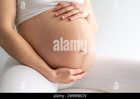 Beschreibung: Mittelteil einer Frau, die auf einem Stuhl sitzt und sanft ihren sehr runden Bauch hält. Seitenwinkelansicht. Weißer Hintergrund. Hell Stockfoto