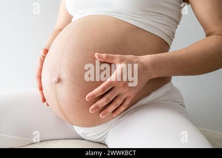 Beschreibung: Mittelteil einer Frau, die auf einem Stuhl sitzt und sanft ihren sehr runden Bauch hält. Seitenwinkelansicht. Weißer Hintergrund. Leuchtend Stockfoto