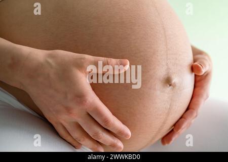 Beschreibung: Nahaufnahme einer Mutter, die sitzt und sanft ihre sehr runde Schwangere hält. Seitenwinkelansicht. Grüner Hintergrund. Brillante Aufnahme. Stockfoto