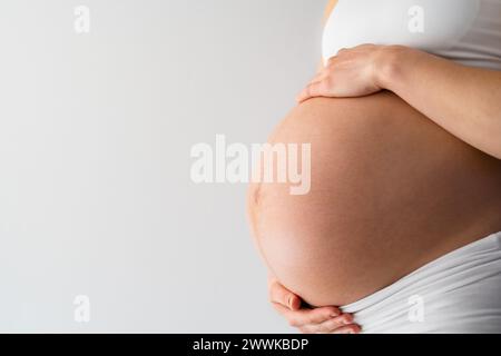 Beschreibung: Mittelteil einer nicht erkennbaren stehenden Mutter, die sanft ihren sehr runden Bauch hält. Seitenwinkelansicht. Weißer Hintergrund. Leuchtend Stockfoto